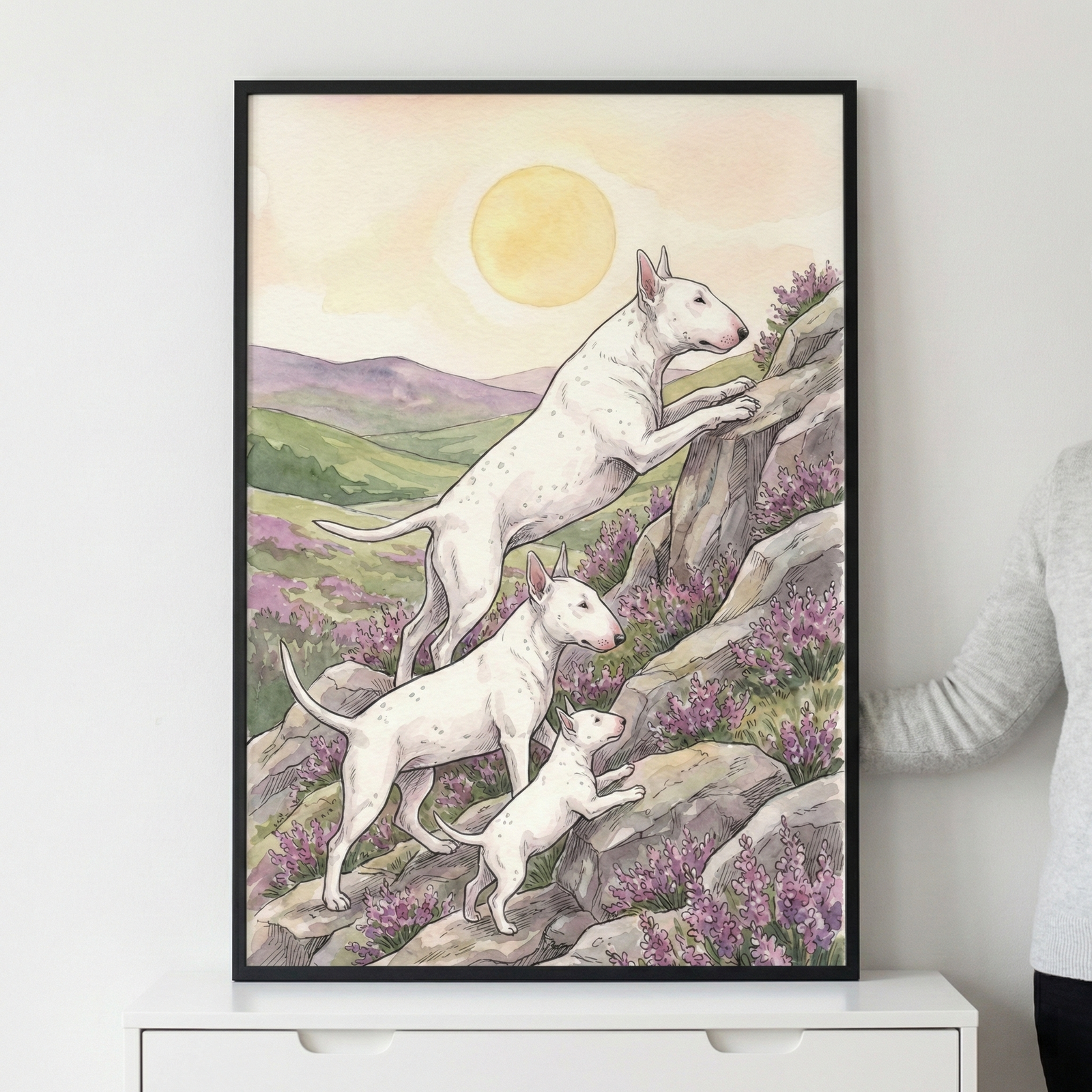 A vertical watercolor-style illustration in a black frame depicts three white dogs with distinctive egg-shaped heads and pointed ears on a rocky hillside. The largest dog at the top reaches its paws up a stone, followed by a medium-sized dog and a small puppy climbing below. They are surrounded by purple flowers blooming among grey rocks. In the background, soft green and purple hills roll beneath a large, pale yellow sun. The framed artwork rests on a white dresser.