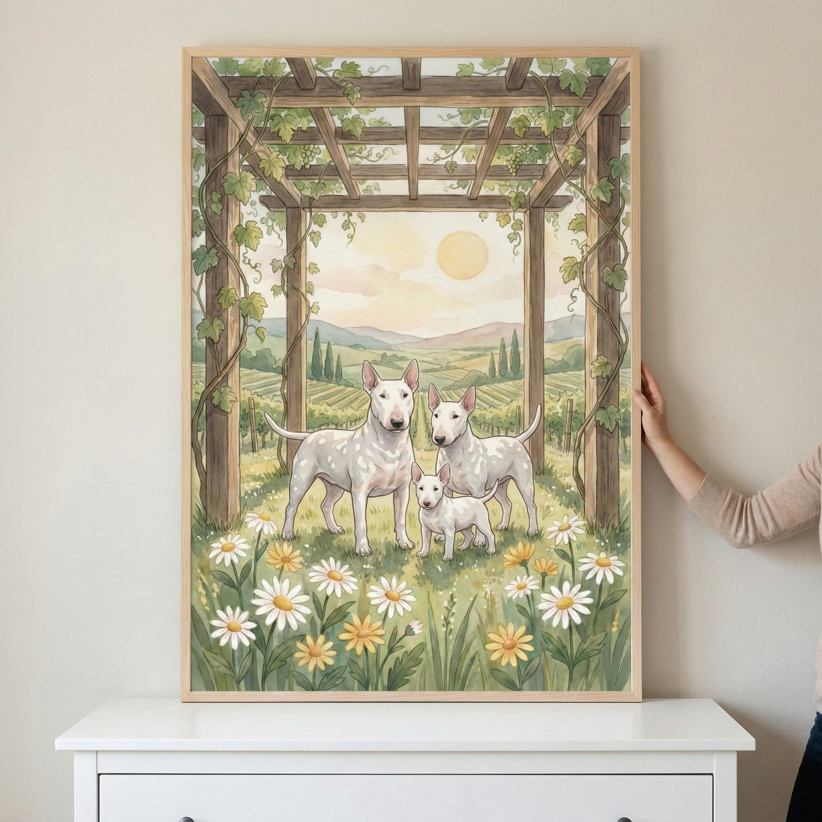A framed illustration depicts a family of three white Bull Terriers: two adults and a puppy. They stand in a grassy field filled with white and yellow daisies. Above them is a wooden pergola with climbing vines. The background shows a peaceful landscape of rolling hills and vineyards under a soft, golden sun. The artwork, shown in a light wood frame, is held against a neutral wall above a white dresser. The style is illustrative with soft colors and delicate textures.