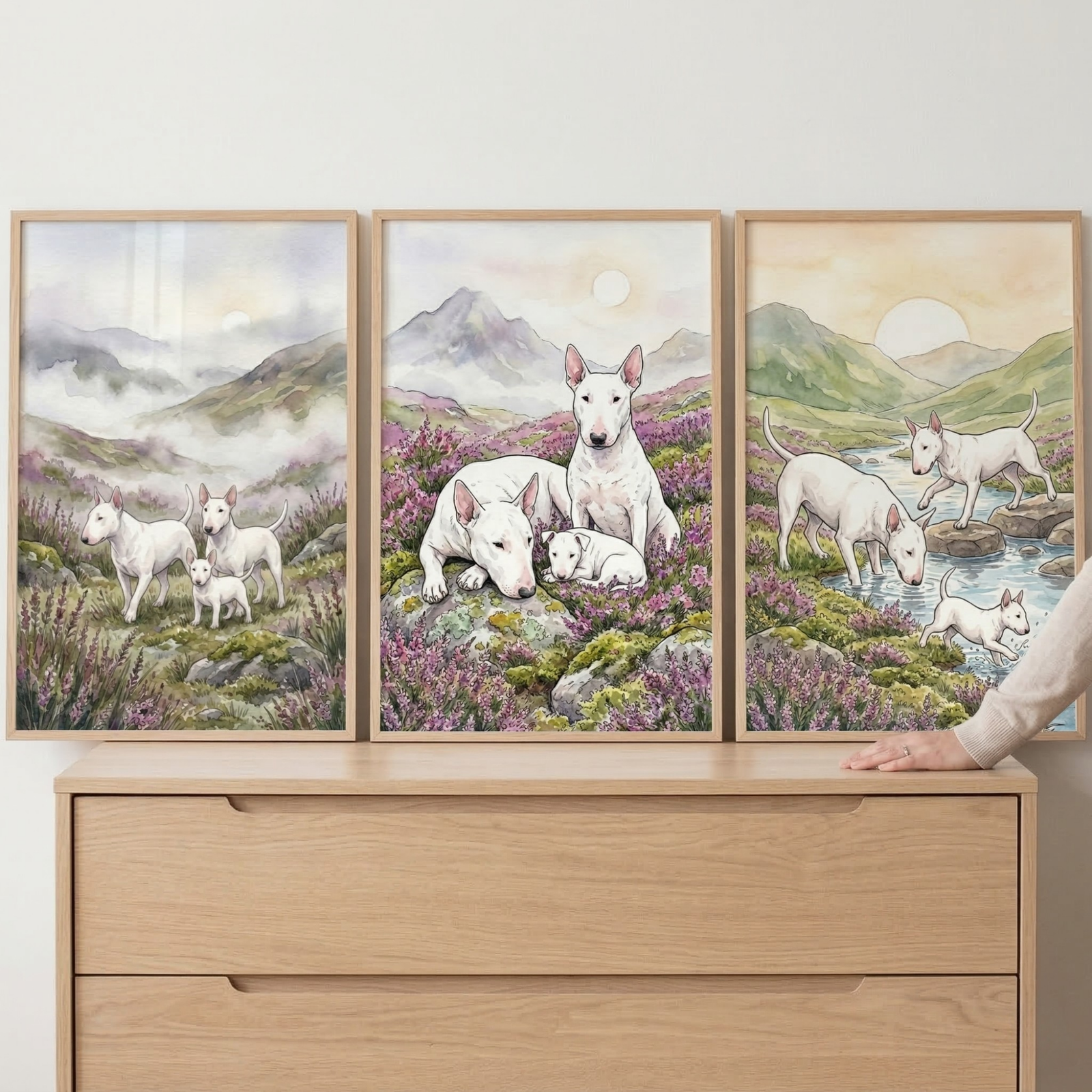 Three framed watercolor prints rest on a wooden dresser, each showing white bull terriers in a soft mountain landscape filled with purple wildflowers. The scenes feature the dogs standing together in misty hills, resting with a puppy among blooming heather, and exploring a shallow stream at sunrise, with a person’s hand visible at the edge of the dresser.