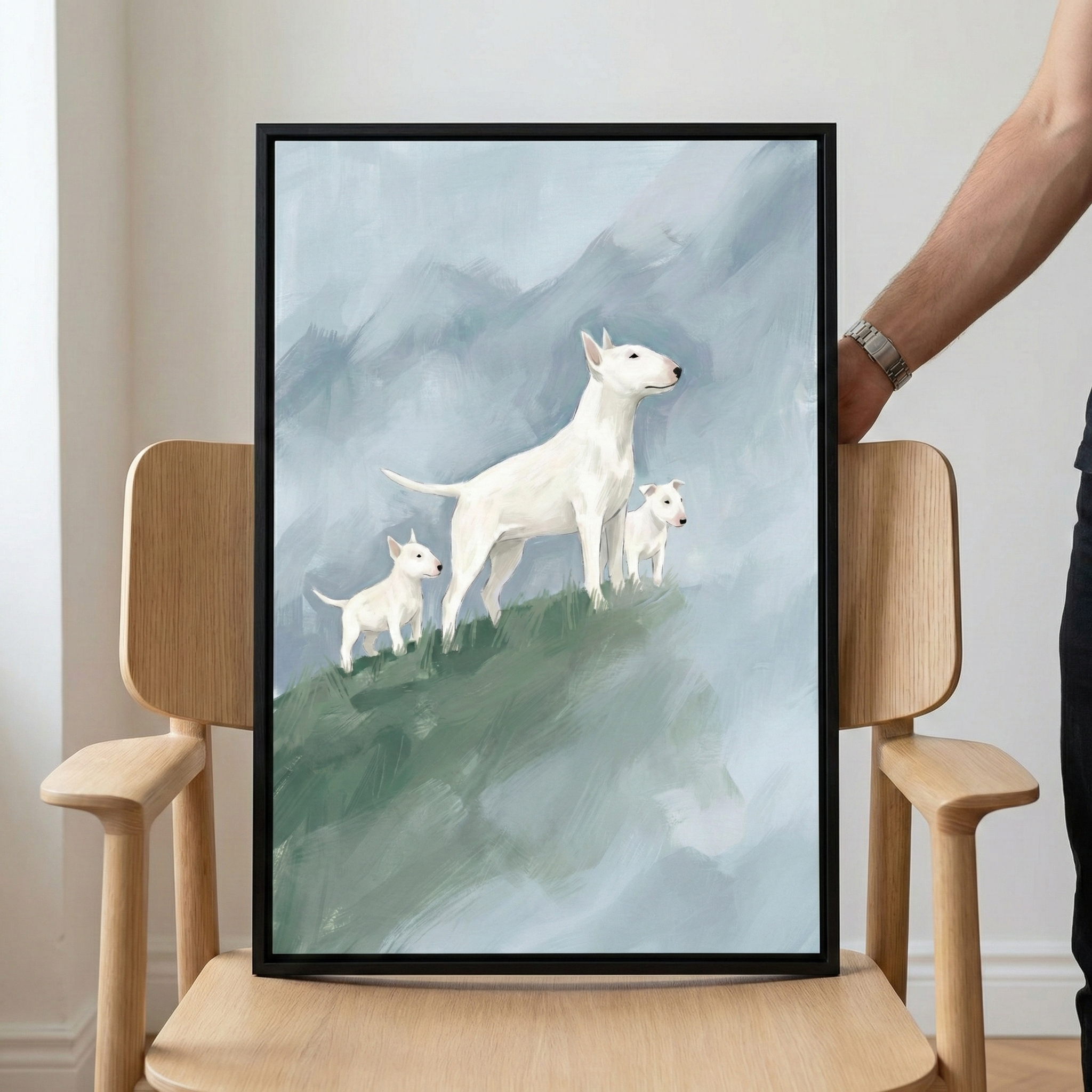 A framed digital illustration features three white dogs with pointed ears and distinctive elongated snouts. An adult dog stands centrally on a textured green slope, accompanied by two smaller puppies. The background consists of broad, atmospheric strokes of light blue and grey, evoking a misty mountainous landscape. The artwork is held in a slim black frame, resting on a light-toned wooden chair, with a person's hand and arm visible on the right side of the frame.