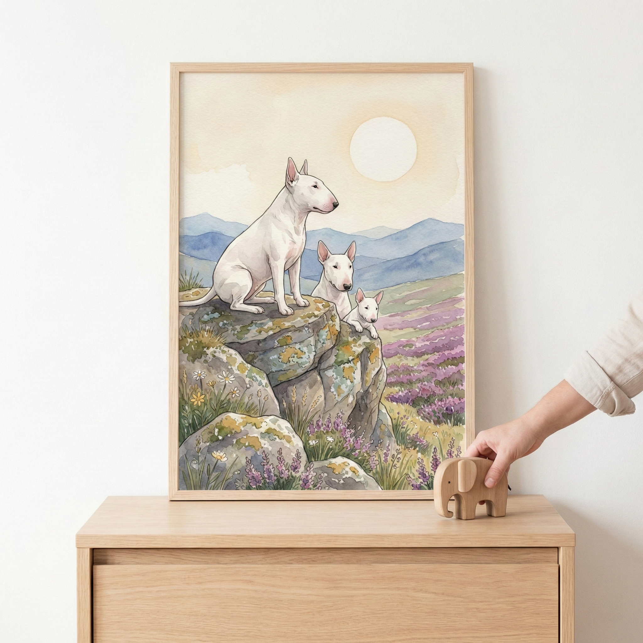 A framed watercolor-style illustration shows three white dogs with distinctive pointed ears and long faces. The largest dog sits atop a rocky outcrop, overlooking a field of purple flowers and distant blue hills. Two smaller dogs peek from behind the stones. A large, soft sun glows in the creamy sky. The artwork is in a light wood frame on a matching dresser, where a hand is placing a small wooden elephant toy. The atmosphere is calm and serene.
