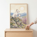 A framed watercolor-style illustration shows three white dogs with distinctive pointed ears and long faces. The largest dog sits atop a rocky outcrop, overlooking a field of purple flowers and distant blue hills. Two smaller dogs peek from behind the stones. A large, soft sun glows in the creamy sky. The artwork is in a light wood frame on a matching dresser, where a hand is placing a small wooden elephant toy. The atmosphere is calm and serene.