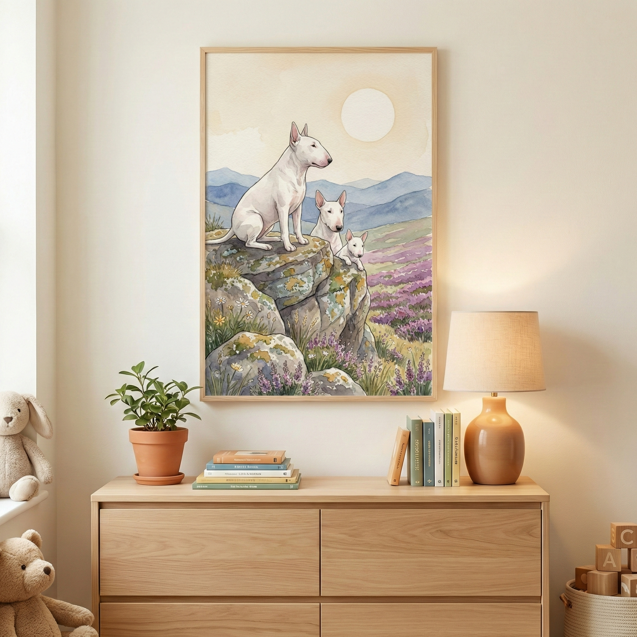 A framed watercolor-style illustration of three white bull terriers—one adult and two smaller puppies—perched on a rocky cliffside. The background features a rolling landscape of purple fields and blue mountains beneath a large pale sun. The frame hangs on a neutral wall above a light wood dresser. On the dresser sits a small potted plant, a stack of books, and a warm lamp. A plush bunny and teddy bear sit on the left, and wooden blocks are visible to the right.