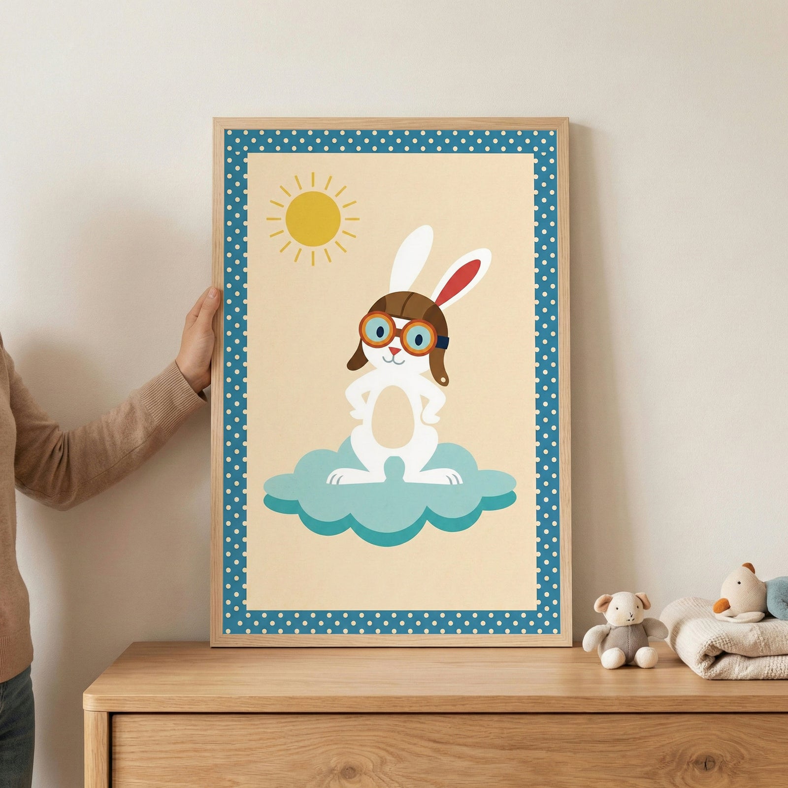 A person's hand holds a framed piece of wall art featuring a white cartoon rabbit dressed as an aviator. The rabbit wears a brown pilot's cap and goggles, standing on a turquoise cloud under a yellow sun. The artwork is set within a light-colored wooden frame and is held above a wooden dresser that displays a small stuffed bear and folded linens.