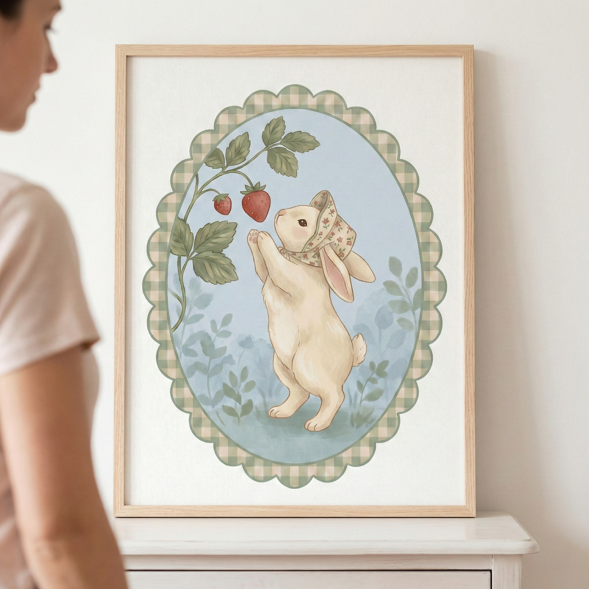 This framed artwork features a charming illustration of a cream-colored rabbit in a whimsical style. The rabbit is depicted standing on its hind legs and wearing a small bonnet with a delicate floral print. It reaches upward with its paws toward a green vine that holds two ripe, red strawberries. 


