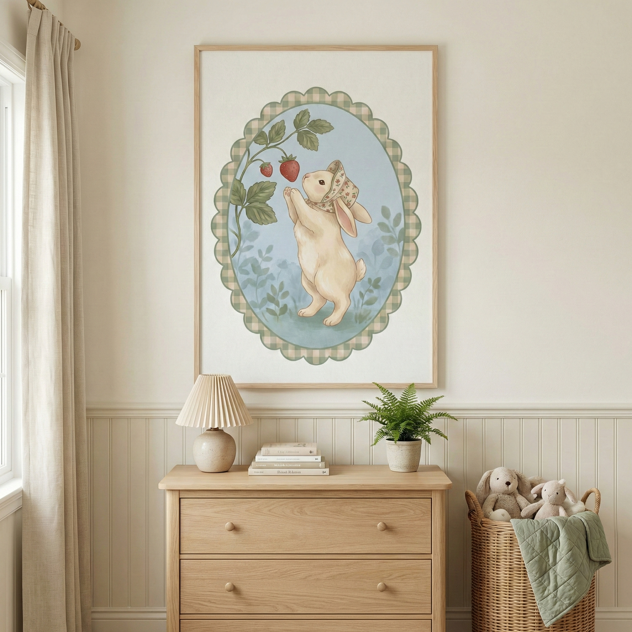 A framed illustration hangs on a light-colored wall above a wooden dresser. The artwork features a cream-colored bunny wearing a floral bonnet, standing on its hind legs to reach for strawberries on a vine. This scene is enclosed in an oval with a green checkered scalloped border. On the dresser sits a pleated lamp, a small stack of books, and a potted plant. To the right, a wicker basket holds plush toy animals and a green blanket. Soft curtains hang by a window on the left.