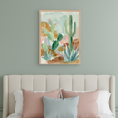 Bedroom with a beige upholstered headboard and pink and blue pillows. A framed watercolor painting of desert cacti and agave plants in light greens, blues, browns, and pinks hangs centered above the headboard.