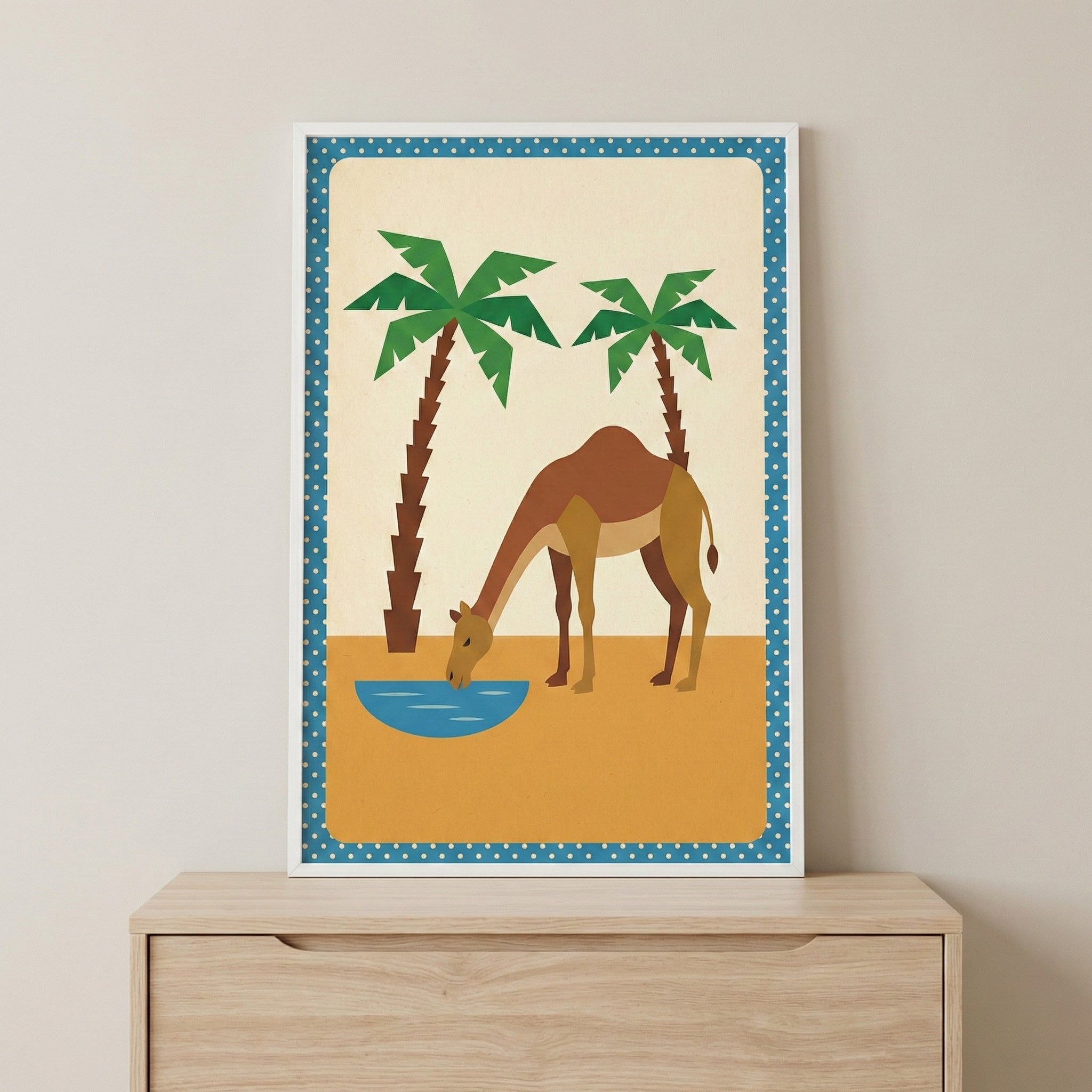 A framed graphic illustration of a brown camel drinking from a small blue pool in a desert setting. Two stylized green palm trees stand behind the camel against a cream-colored background. The ground is represented by a solid mustard-yellow field. The artwork features a blue border with white polka dots and is held in a thin white frame, resting on top of a light-colored wooden dresser against a neutral wall.
