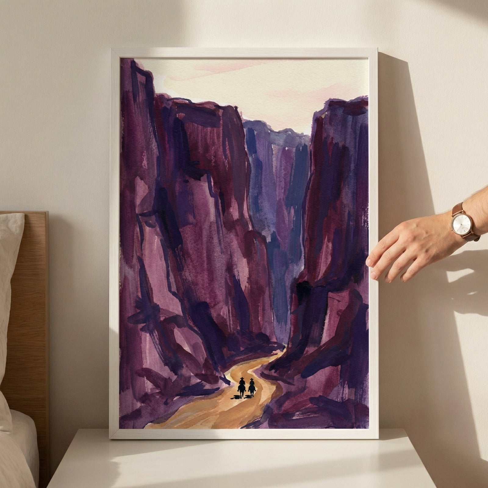 Framed painting of two small riders on horseback following a winding path through towering canyon walls in deep shades of purple and red. The stylized western landscape evokes a dramatic desert scene with a sense of scale and solitude.