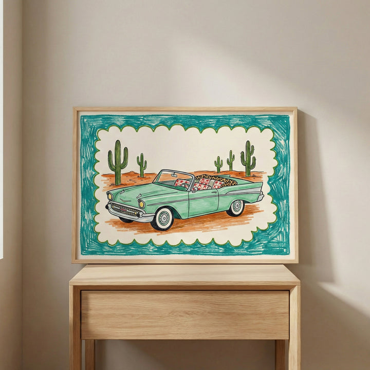 Framed artwork of a green vintage convertible car driving through a desert with tall cacti, bordered by a teal and scalloped frame, displayed on a light wood table against a neutral wall.