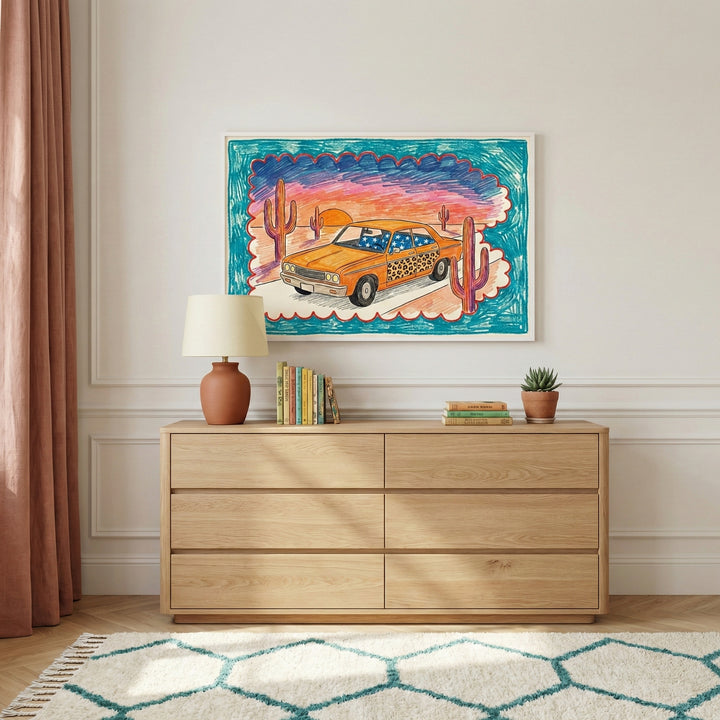 Framed drawing of a desert car scene hanging above a light wood dresser in a modern interior. The illustration features a sunset backdrop with cacti and an orange car, blending playful patterns like stars and leopard print.