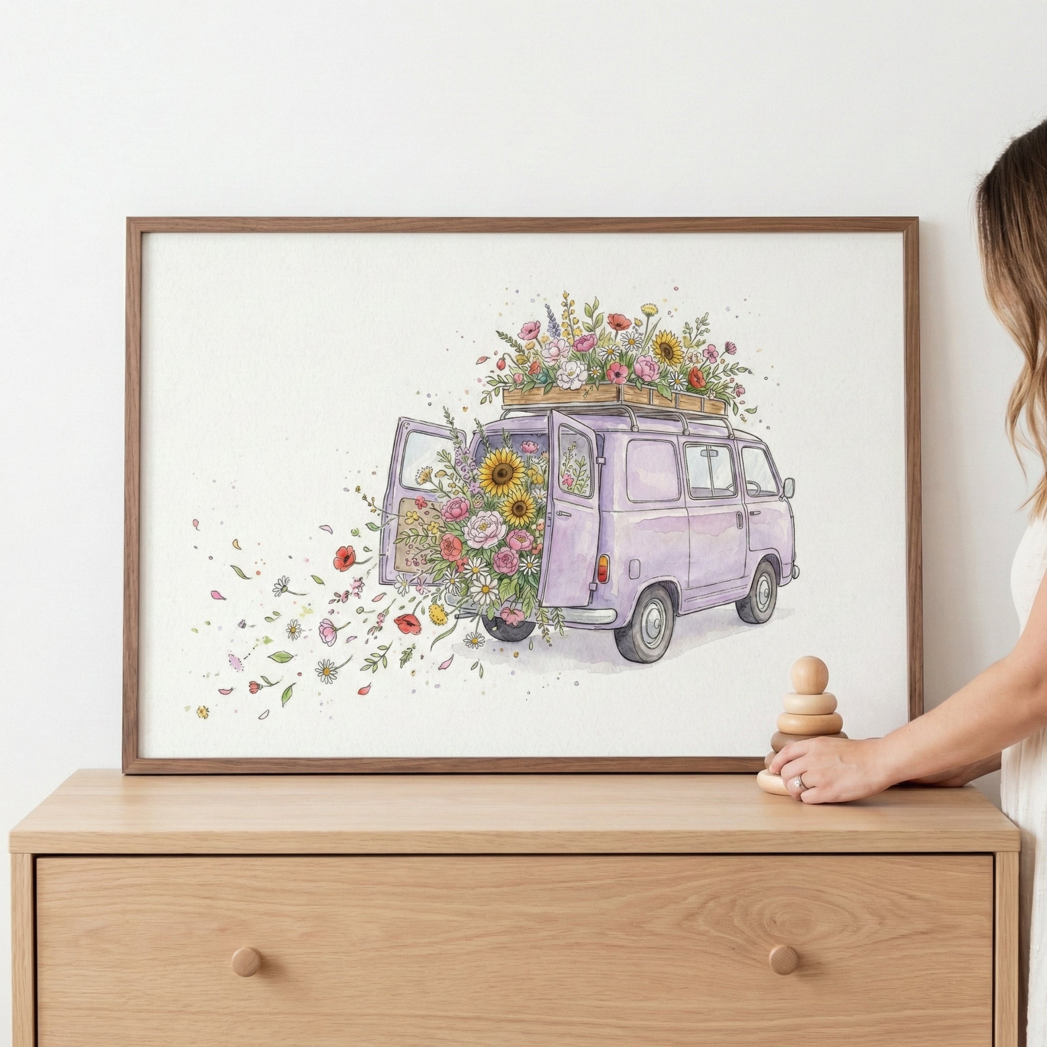 A framed watercolor painting features a lavender-colored vintage van overflowing with flowers from the back and roof, with petals floating outward in a whimsical swirl. The artwork rests on a wooden dresser beside a woman’s hand holding stacked wooden toys.