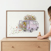 A framed watercolor painting features a lavender-colored vintage van overflowing with flowers from the back and roof, with petals floating outward in a whimsical swirl. The artwork rests on a wooden dresser beside a woman’s hand holding stacked wooden toys.