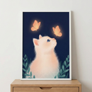A framed digital illustration features a soft, fluffy white kitten looking upward at two glowing orange butterflies against a dark navy blue background. The kitten has large blue eyes and is surrounded by stylized green leaves at the bottom of the frame. The butterflies emit a warm light that highlights the kitten's face. The artwork is held in a minimalist white frame, resting on a light-colored wooden cabinet against a plain white wall.
