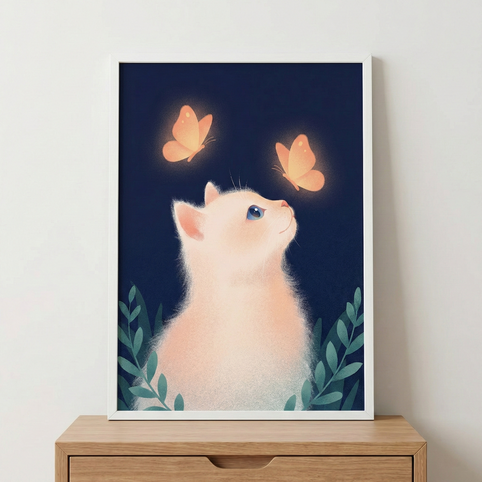 A framed digital illustration features a soft, fluffy white kitten looking upward at two glowing orange butterflies against a dark navy blue background. The kitten has large blue eyes and is surrounded by stylized green leaves at the bottom of the frame. The butterflies emit a warm light that highlights the kitten's face. The artwork is held in a minimalist white frame, resting on a light-colored wooden cabinet against a plain white wall.