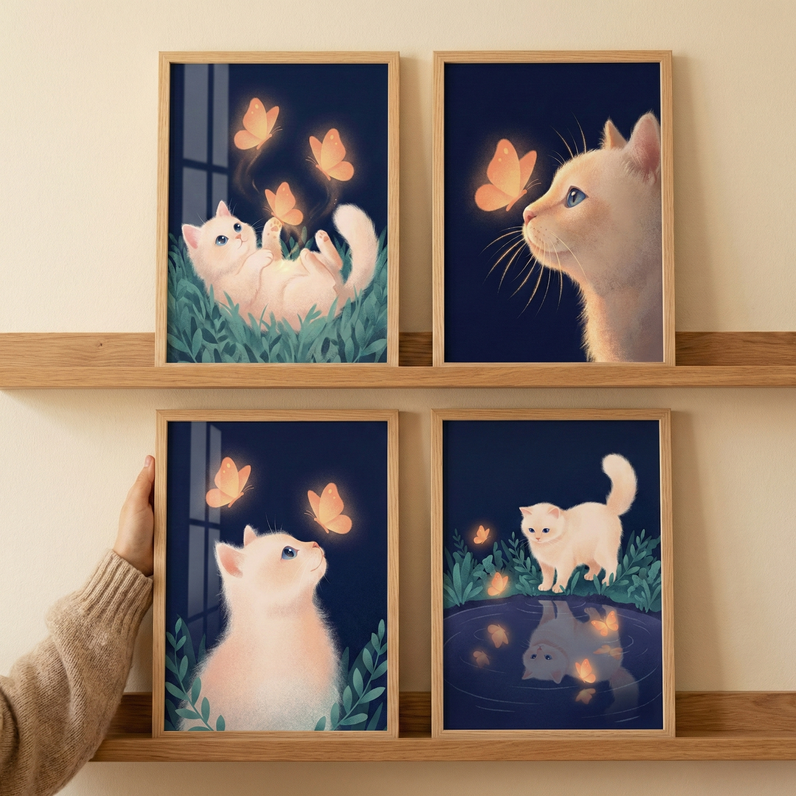 A set of four wooden frames arranged in a two-by-two grid on two light-colored wooden shelves. A person's hand is seen touching the bottom-left frame. Each frame contains a whimsical illustration featuring a soft, light-colored, fluffy cat and glowing orange butterflies against a dark blue background. The top-left illustration shows the cat lying in green grass, looking at several glowing butterflies. The top-right shows a close-up of the cat's face as it watches a butterfly. 