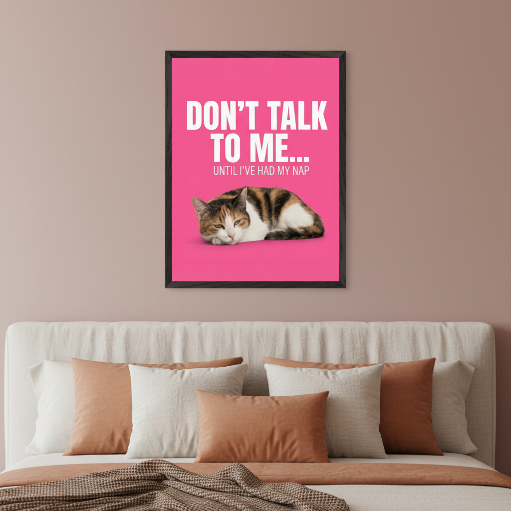 Modern bedroom with a bed featuring a light headboard and brown/white pillows. A black-framed pink poster with white text "DON'T TALK TO ME... UNTIL I'VE HAD MY NAP" and a sleeping calico cat is centered above the bed.