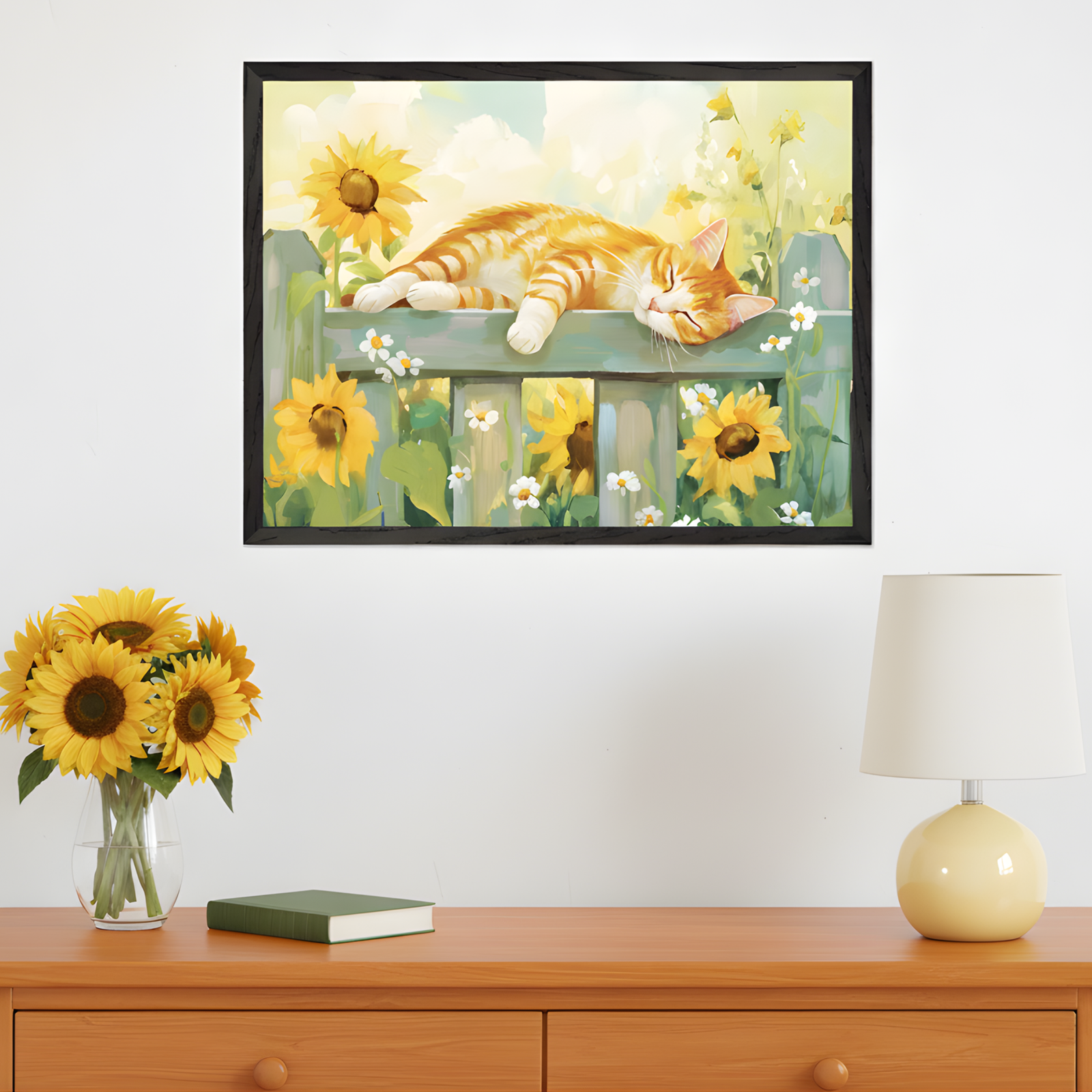 Framed artwork of an orange tabby cat peacefully napping on a wooden fence surrounded by blooming sunflowers and white daisies. The artwork hangs above a wooden dresser with a vase of fresh sunflowers, a closed book, and a cream-colored lamp, adding a cozy and cheerful touch to the space.