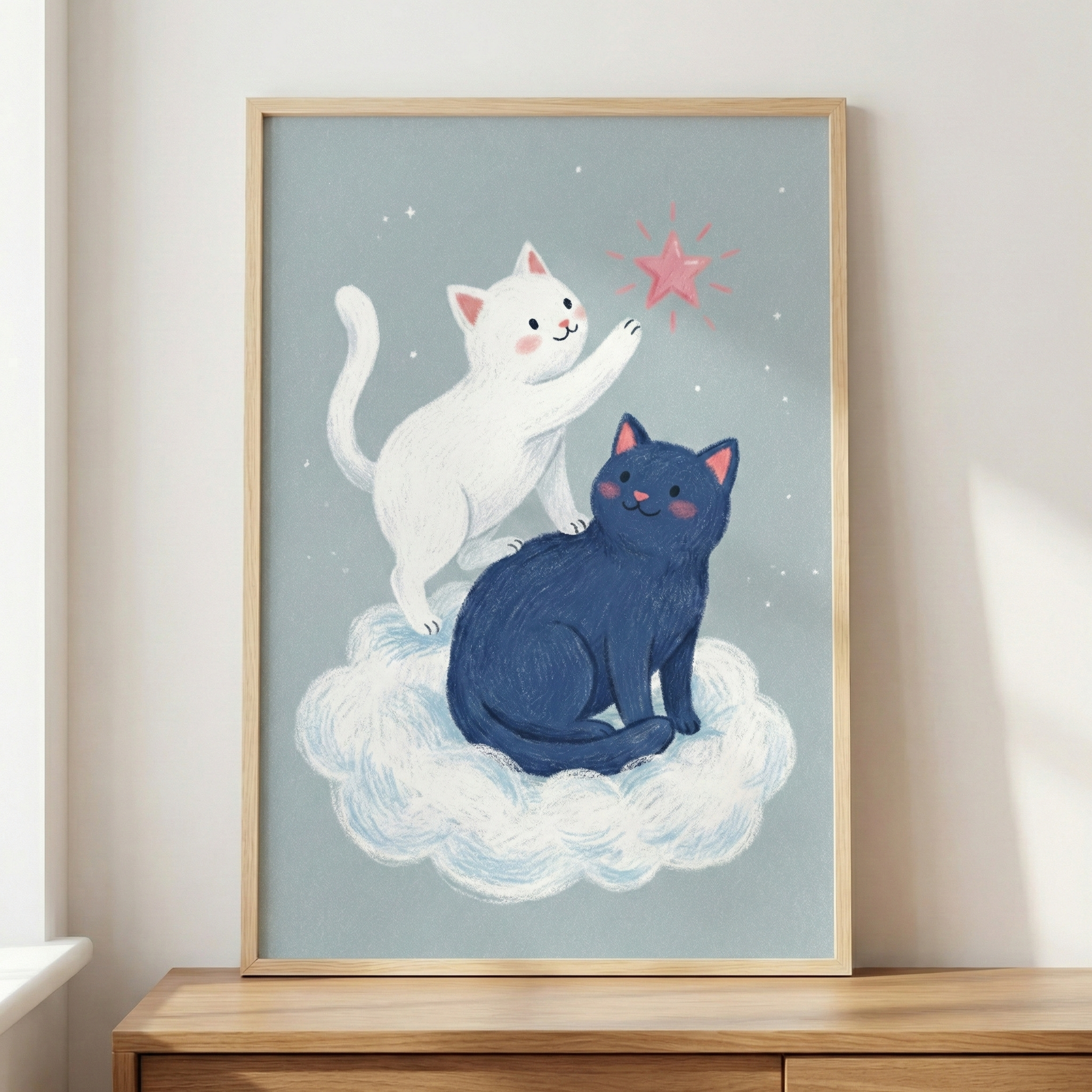 This framed piece of art shows two whimsical cats atop a soft, white cloud. A small white cat stands on its hind legs, reaching a paw toward a sparkling pink star in a muted blue sky filled with tiny specks. Below it sits a larger, dark blue cat with a gentle expression. The illustration has a textured, hand-drawn look and is held within a simple light wood frame, resting on a wooden tabletop against a neutral background.