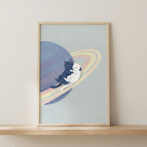 A framed illustration shows two cute cartoon cats, one black and one white, hugging each other while sitting on the multi-colored rings of a large, textured planet. The planet is colored in shades of purple and blue, and the rings feature soft pastel tones of pink, yellow, and blue. This whimsical artwork is set against a solid light-blue background. The piece is held in a simple, light-toned wooden frame and is displayed on a wooden shelf in front of a neutral-colored wall.