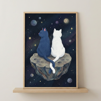 A vertical framed illustration of two cats, one dark blue and one white, sitting side-by-side on a small, cratered asteroid in outer space. The cats are viewed from behind, their long tails gently crossed. The dark cosmic background is dotted with stars and colorful, distant planets. The artwork is set in a light wood frame, resting on a wooden shelf against a neutral-toned wall.