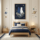 A cozy, space-themed bedroom features a light wood bed frame dressed in dark blue and cream-colored bedding. Above the bed, a large framed illustration depicts a dark blue cat and a white cat sitting side-by-side on a cratered asteroid, looking out at a starry, deep-blue cosmos. The off-white wall on either side of the frame is decorated with decals of planets and stars in a muted color palette. 