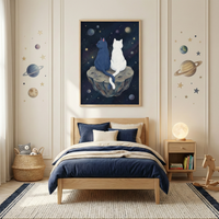 A cozy, space-themed bedroom features a light wood bed frame dressed in dark blue and cream-colored bedding. Above the bed, a large framed illustration depicts a dark blue cat and a white cat sitting side-by-side on a cratered asteroid, looking out at a starry, deep-blue cosmos. The off-white wall on either side of the frame is decorated with decals of planets and stars in a muted color palette. 