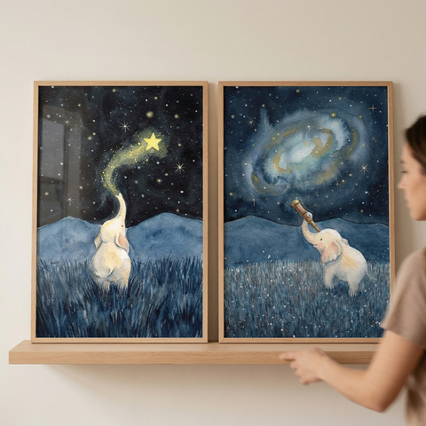 Two framed watercolor-style illustrations rest on a light wood shelf against a neutral wall. On the left, a small white elephant gazes at a starry night sky, with a brilliant yellow star and its glowing trail appearing to come from its trunk. On the right, the same elephant uses a telescope to view a large, swirling galaxy. Both pieces feature blue mountains and a grassy foreground. A person's arm and shoulder are visible as they position the frames.