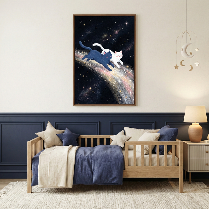 A vertical framed artwork hangs on a light cream wall above a dark blue wainscoted section in a cozy bedroom. The artwork depicts two whimsical cats—one deep blue and one white—leaping along a vibrant, swirling cosmic path of multi-colored stardust and glowing stars against a dark night sky. Below, a light wood toddler bed is dressed in blue and cream bedding with star-shaped pillows. To the right, a warm lamp sits on a small nightstand, beneath a hanging moon-and-stars mobile.