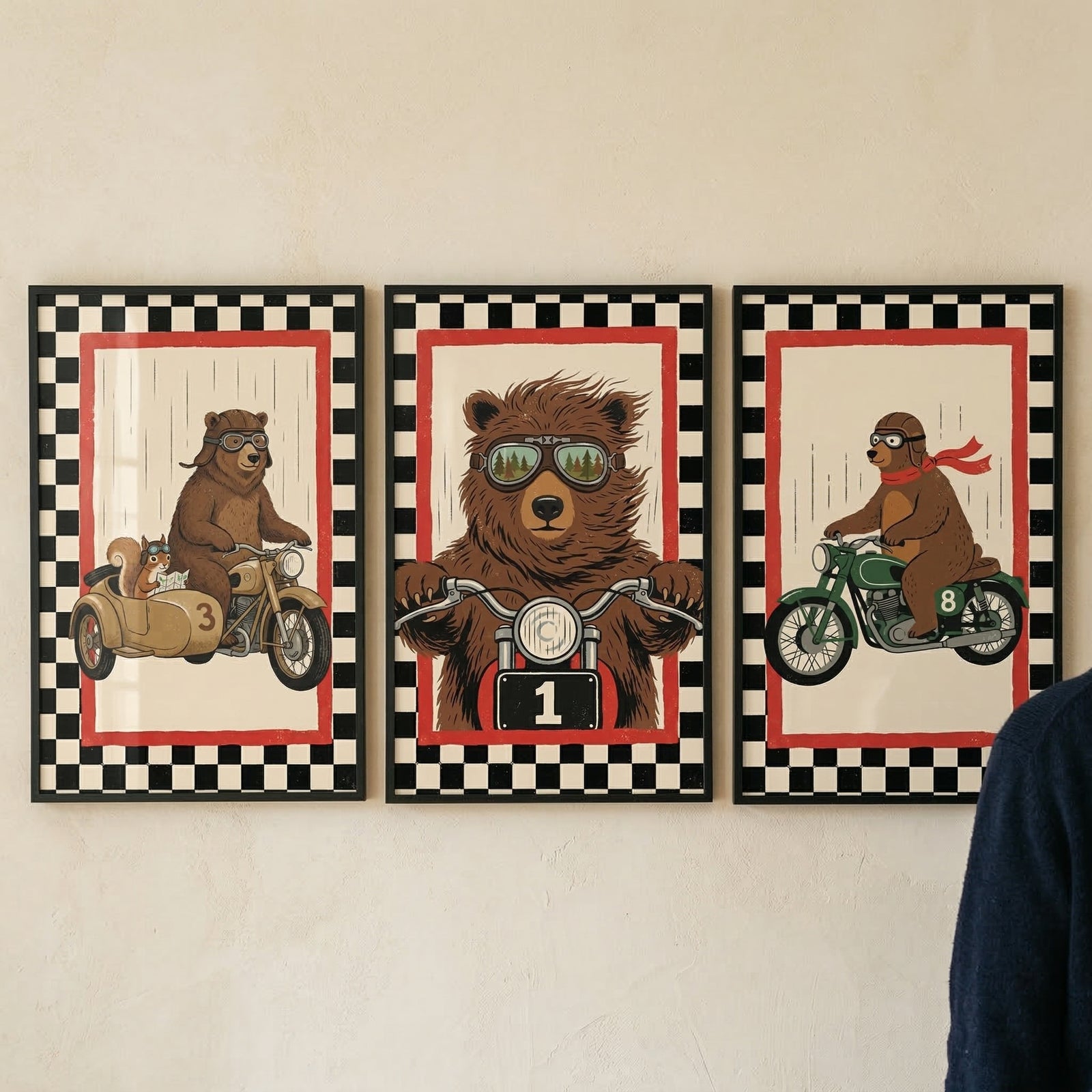 Three framed illustrations are hung side-by-side on a light-colored wall, each featuring a brown bear on a motorcycle. Each piece is set within a black frame and has a border featuring a black and white checkerboard pattern followed by a thin red inner line.

