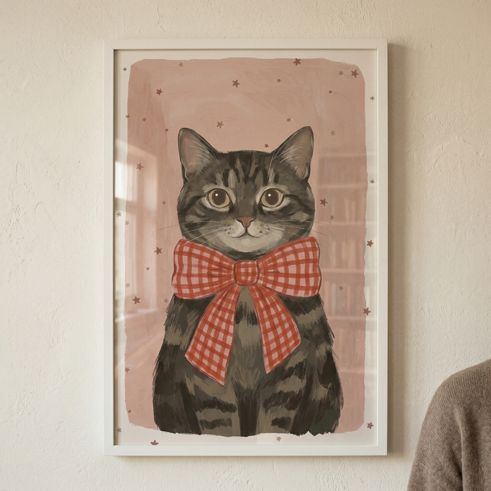 A framed stylized illustration of a brown tabby cat hangs on a light, textured wall. The cat is shown from the front with large, amber-colored eyes and a small pink nose. It wears a large, red and white checkered bow tie around its neck. The artwork's background is a dusty pink with small, dark stars scattered throughout. The piece is held in a simple white frame. A person's shoulder in a brown knit sweater is partially visible on the right edge.