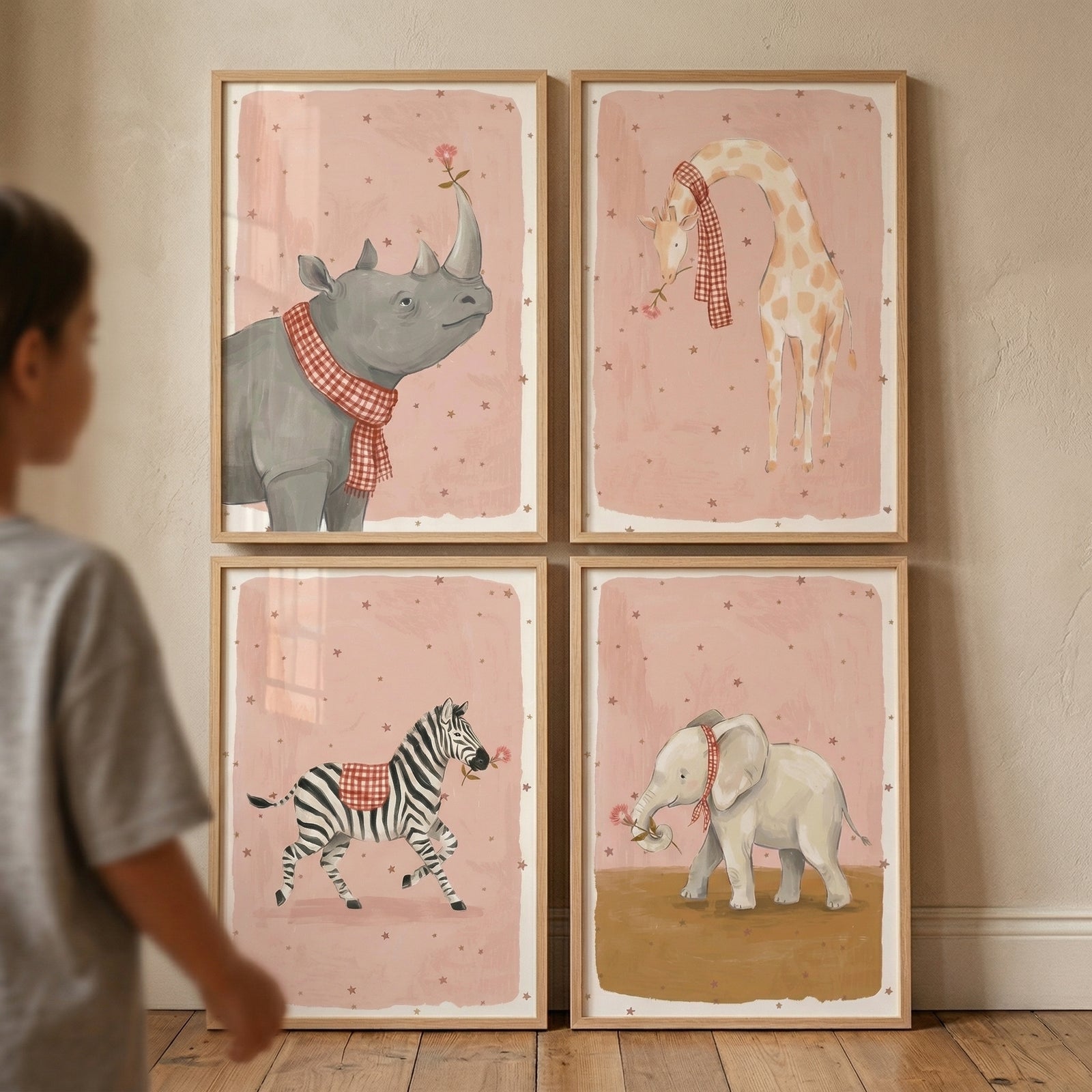 Four rectangular wooden frames are arranged in a square on a light, textured wall. Each frame contains a whimsical illustration of a safari animal against a soft pink background scattered with small brown stars. The top left features a gray rhinoceros wearing a red and white checkered scarf with a small flower on its horn. 