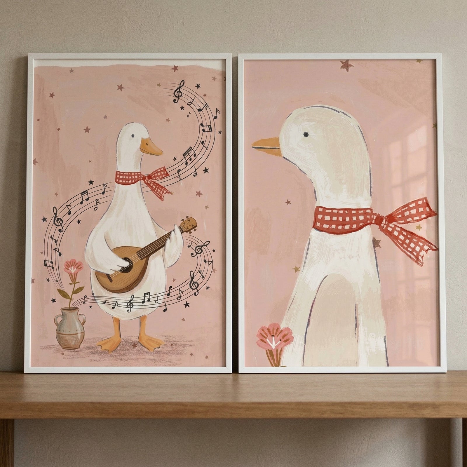Two framed pieces of illustrative art featuring a white waterfowl are displayed side-by-side on a light-colored wooden shelf. Both pieces are set against a soft, dusty pink background adorned with small, delicate stars.
