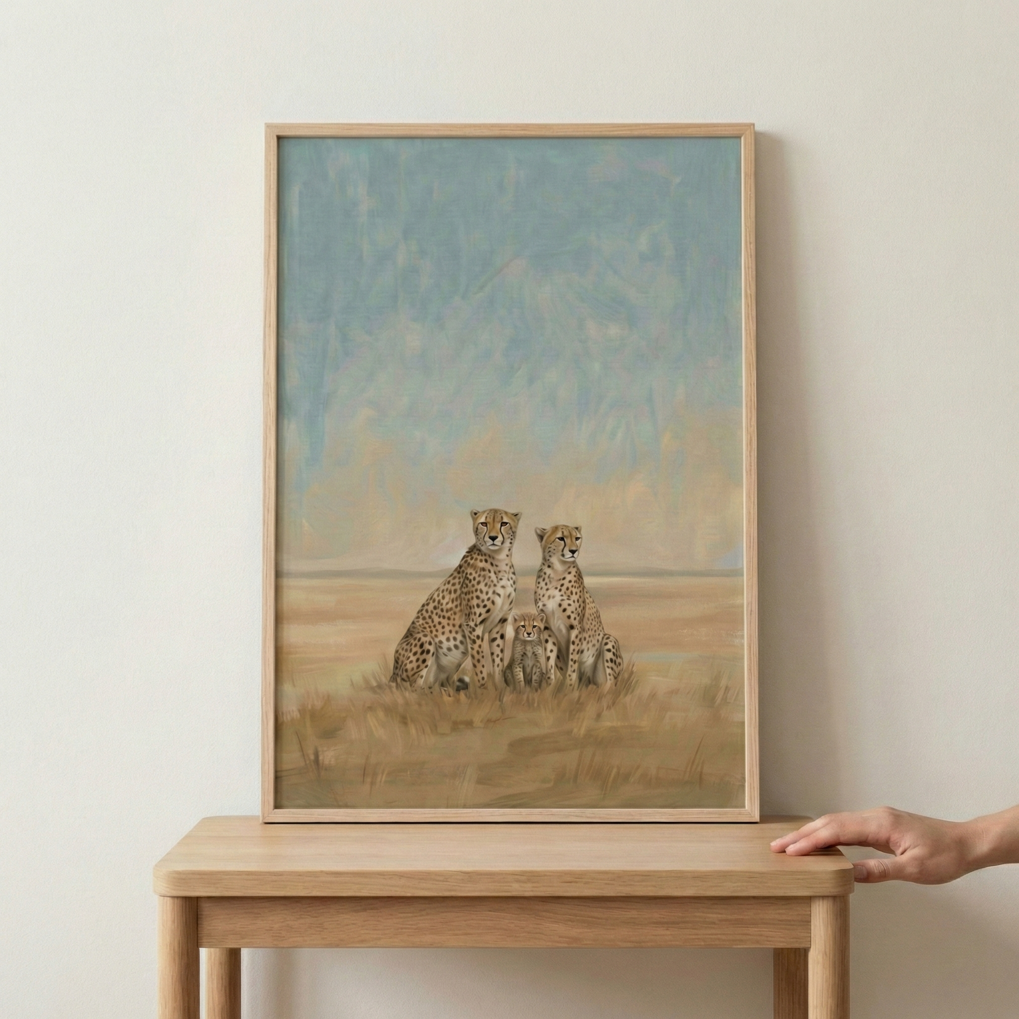 An illustrative piece of artwork featuring a family of cheetahs is shown within a light wood frame. Two adult cheetahs sit side-by-side, with two small cubs nestled between them. The background depicts an expansive savanna landscape under a muted blue sky, rendered with soft brushstrokes and a warm, earthy color palette. The framed image rests on a wooden table, and a hand is visible on the right side, just touching the edge of the tabletop.