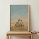 An illustrative piece of artwork featuring a family of cheetahs is shown within a light wood frame. Two adult cheetahs sit side-by-side, with two small cubs nestled between them. The background depicts an expansive savanna landscape under a muted blue sky, rendered with soft brushstrokes and a warm, earthy color palette. The framed image rests on a wooden table, and a hand is visible on the right side, just touching the edge of the tabletop.