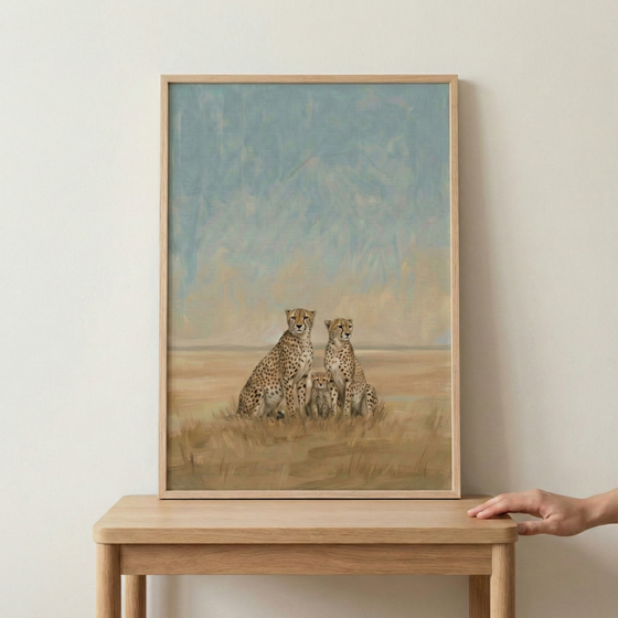 An illustrative piece of artwork featuring a family of cheetahs is shown within a light wood frame. Two adult cheetahs sit side-by-side, with two small cubs nestled between them. The background depicts an expansive savanna landscape under a muted blue sky, rendered with soft brushstrokes and a warm, earthy color palette. The framed image rests on a wooden table, and a hand is visible on the right side, just touching the edge of the tabletop.