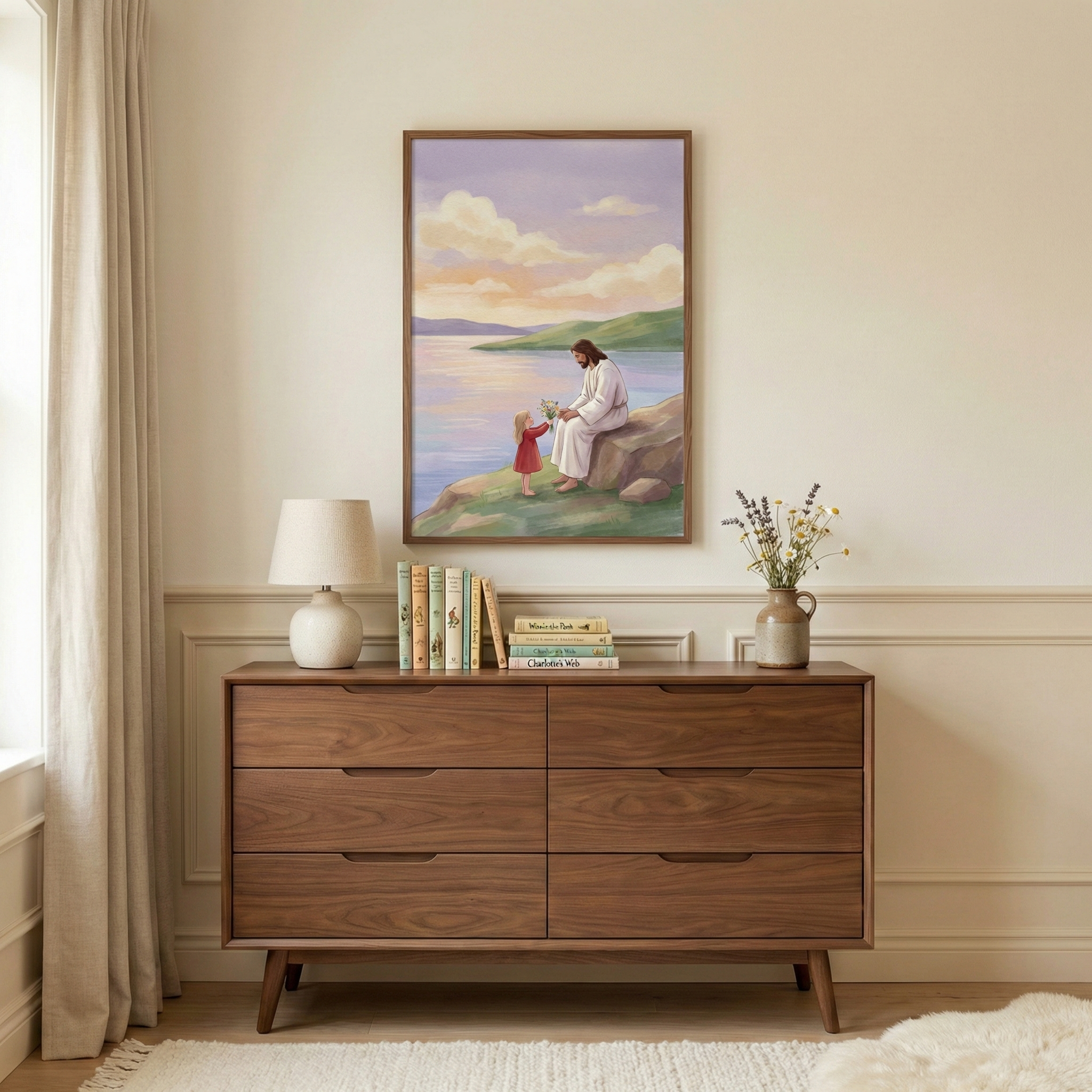 A framed artwork hangs on a beige wall above a mid-century modern style wooden dresser. The artwork depicts Jesus sitting on a rock by water, sharing a moment with a small girl in a red dress under a pastel sky. On the dresser is a white lamp, a collection of books, and a small vase with flowers. A light-colored curtain hangs to the left, and a white rug is at the bottom. The room has a calm, peaceful feel with neutral tones and soft lighting.