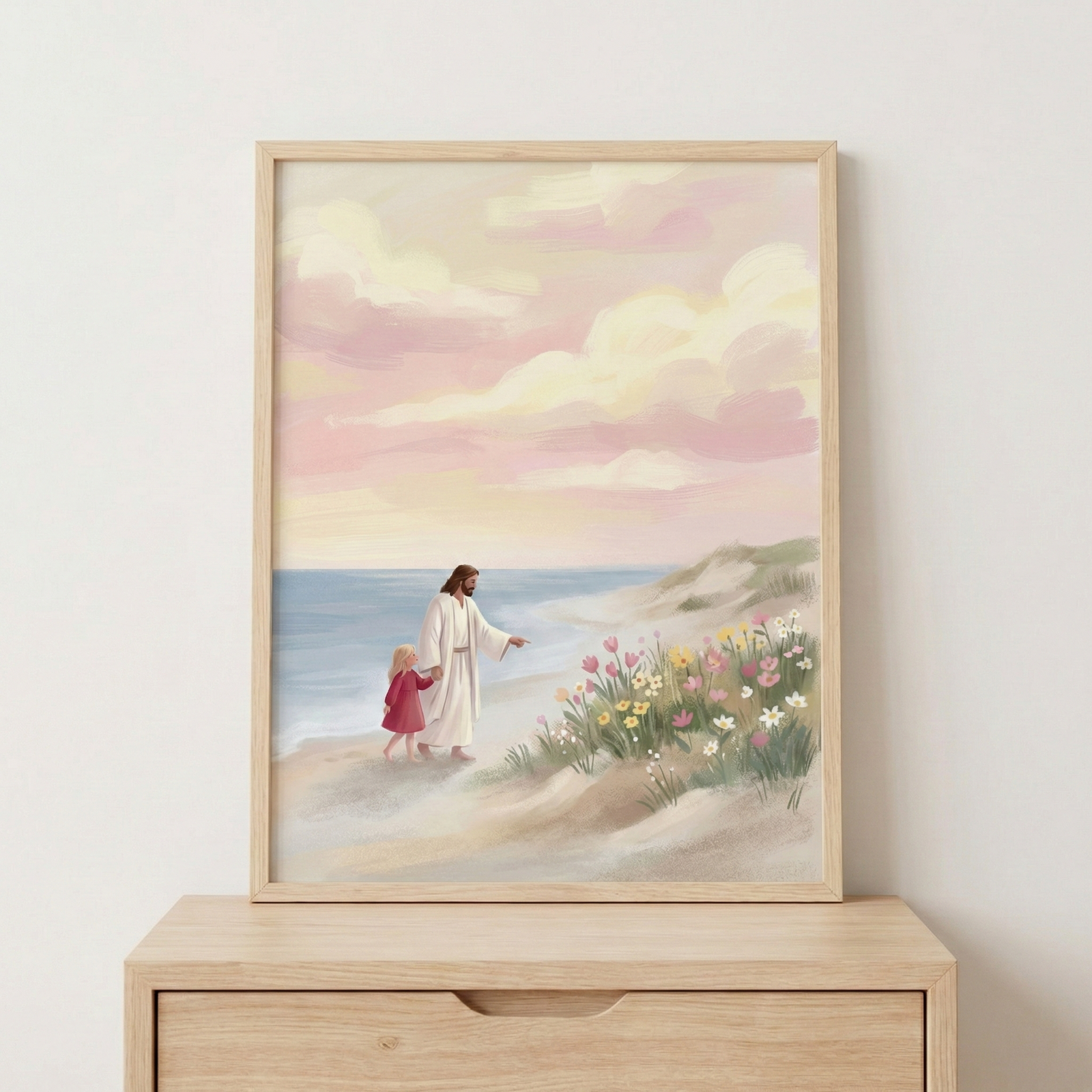 An illustrative artwork sits in a light-colored wood frame on top of a wooden dresser. The image depicts Jesus Christ walking along a sandy beach with a small child. Jesus has long, flowing brown hair and is wearing a simple white robe. He holds the hand of a little girl with blonde hair who is wearing a red dress. With his other hand, Jesus points toward a cluster of colorful wildflowers growing on a grassy dune. The flowers are in shades of pink, yellow, and white.
