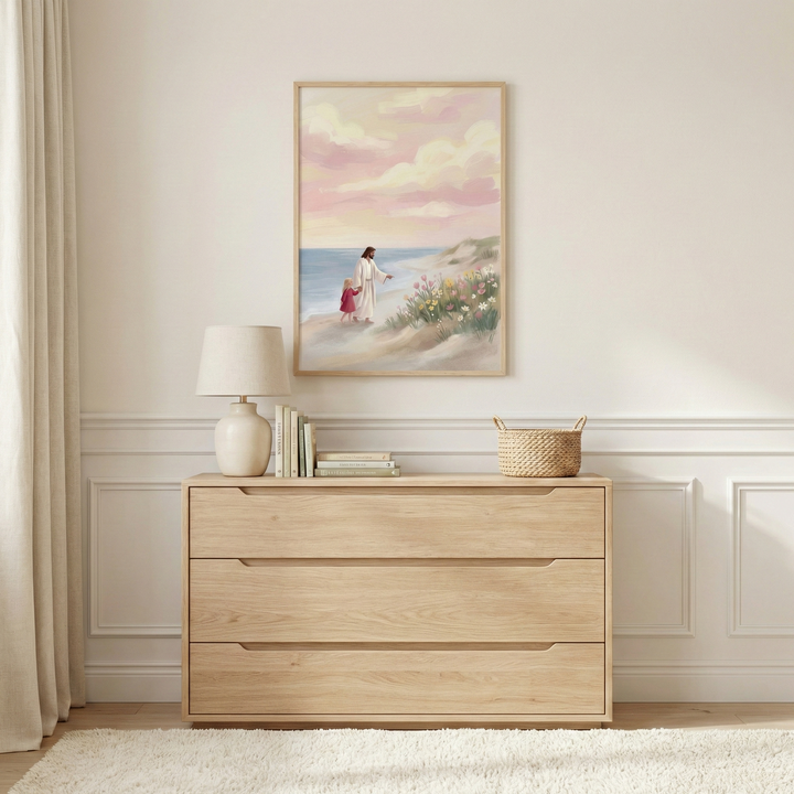 This interior shot showcases a framed piece of art mounted on a light-colored wall above a minimalist wooden dresser. The artwork depicts Jesus walking along a sandy beach with a young child in a red dress, headed toward a flower-covered dune. The background of the art features a calm blue sea under a sky filled with soft pink and white clouds.

