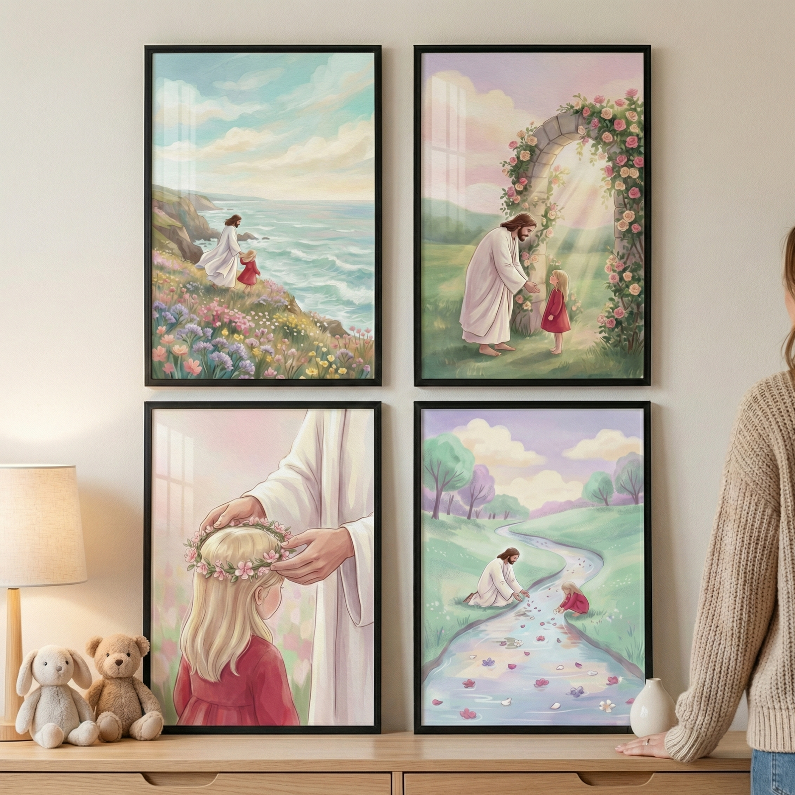 A set of four black-framed illustrations is arranged in a square on a light-colored wall above a wooden dresser. On the dresser, a lamp with a beige shade stands next to two small plush toys, a white rabbit and a brown bear. To the right, the back of a person wearing a beige knit sweater is partially visible. Each of the four illustrations depicts a gentle scene between Jesus and a young blonde girl in a red dress.  