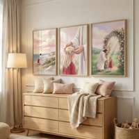 A serene, brightly lit room features a light-colored wood dresser as the focal point. Atop the dresser are several soft, textured pillows in shades of cream and blush pink, along with a cozy cream-colored knit throw draped over the front. Above the dresser, three light-wood framed illustrations are hung in a row. The first shows Jesus Christ walking along a beach with a young child in a red dress under a soft pink sky. 