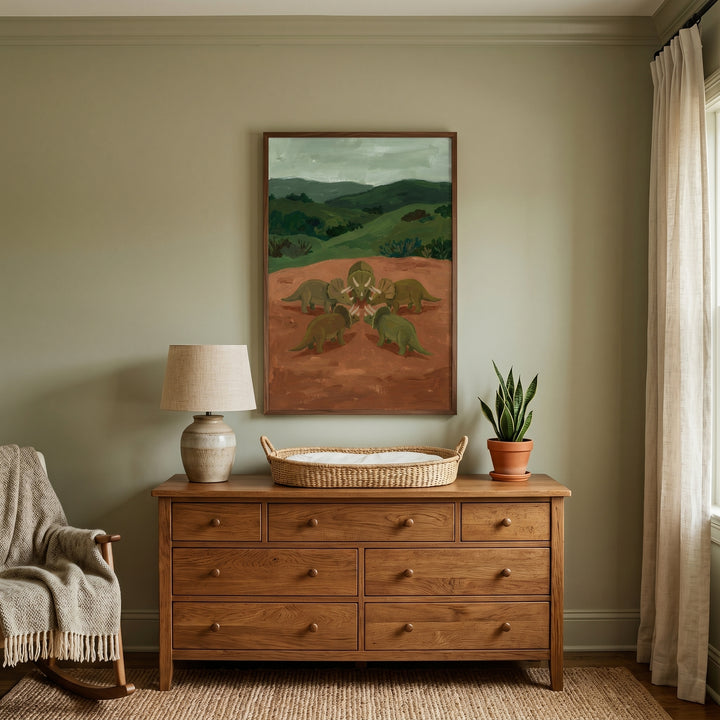 Wooden dresser below circle five green dinosaur wall art in a sage green room with a lamp, plant, and woven basket.