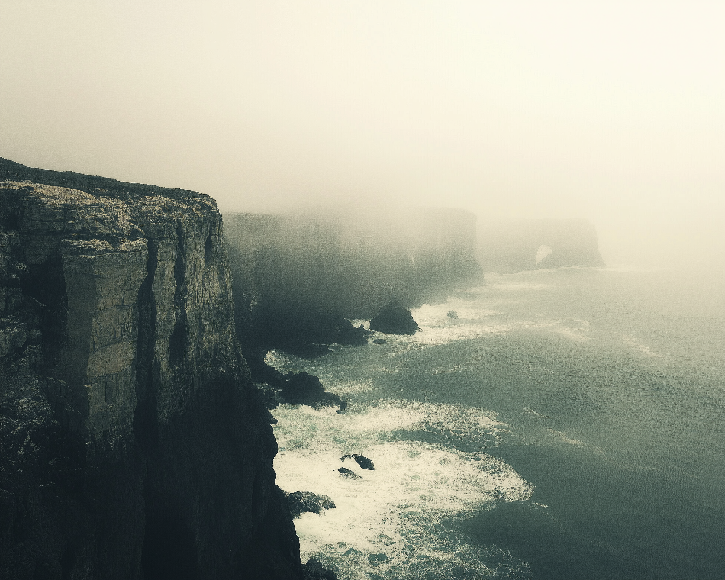 Steep sea cliffs rise sharply above crashing waves, disappearing into thick coastal fog with a natural rock arch faintly visible in the distance.
