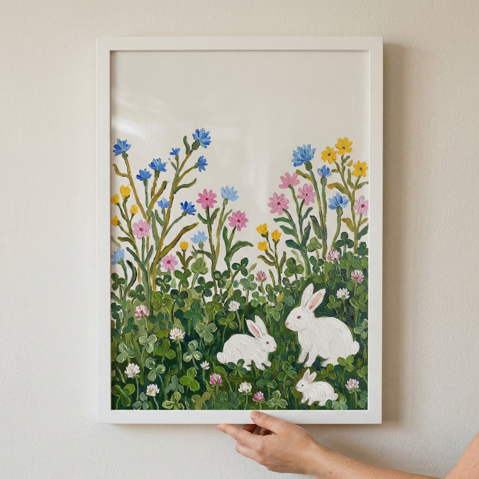 This illustrative artwork showcases three white rabbits in a lush meadow of green clover and colorful wildflowers. The floral arrangement includes delicate blooms in blue, pink, and yellow hues. A large rabbit and two smaller ones are scattered across the verdant scene. 