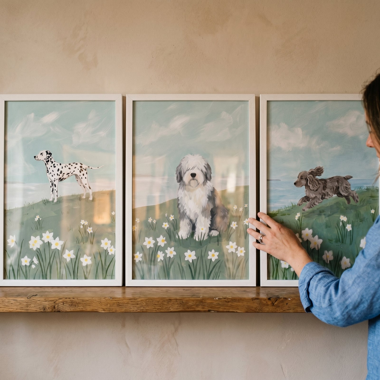 A hand touches a coastal flower field dog wall art set of 3 featuring a dalmatian, sheepdog, and spaniel on a shelf.