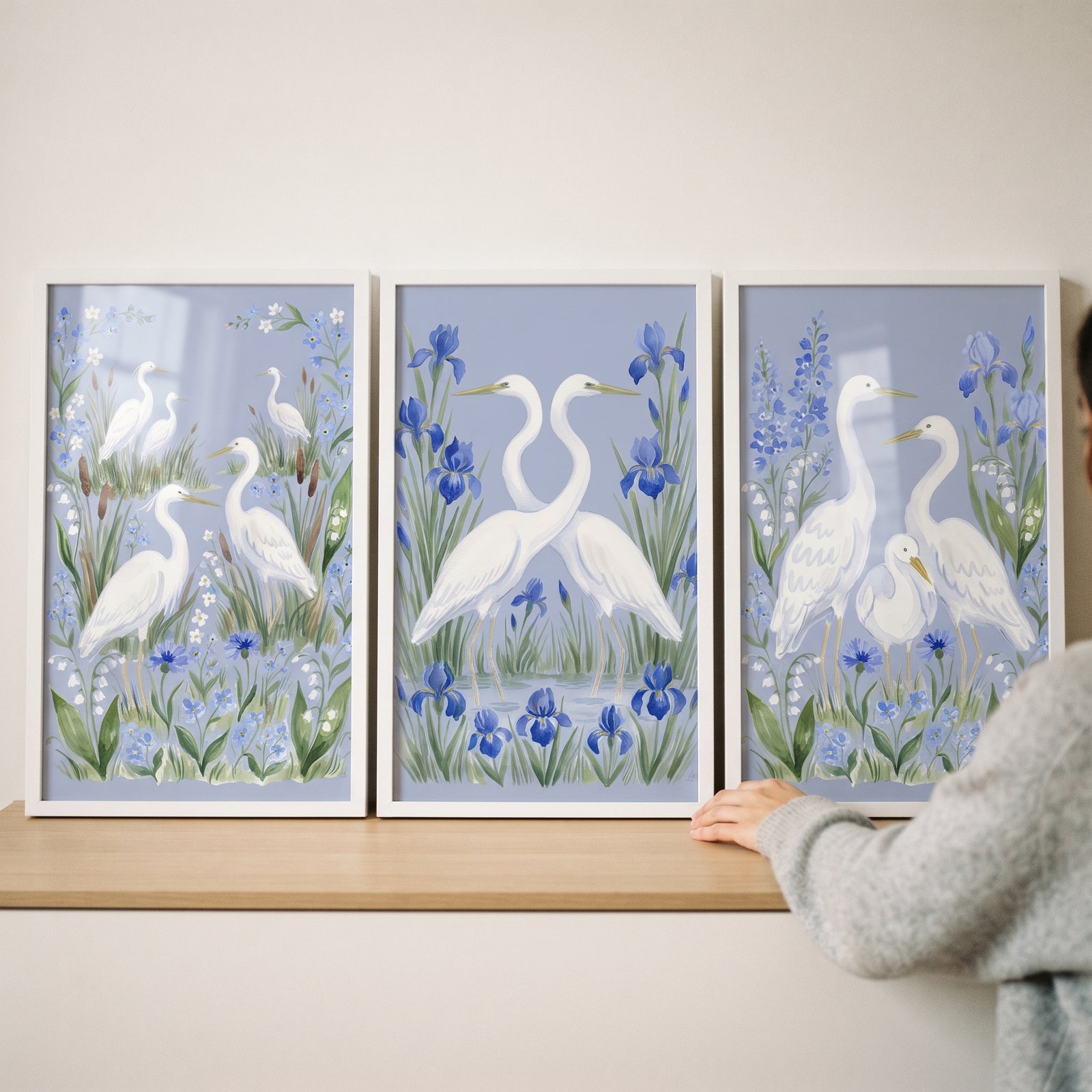 Coastal garden white egret bird wall art set of 3 shows white birds among blue flowers in frames on a wooden shelf.