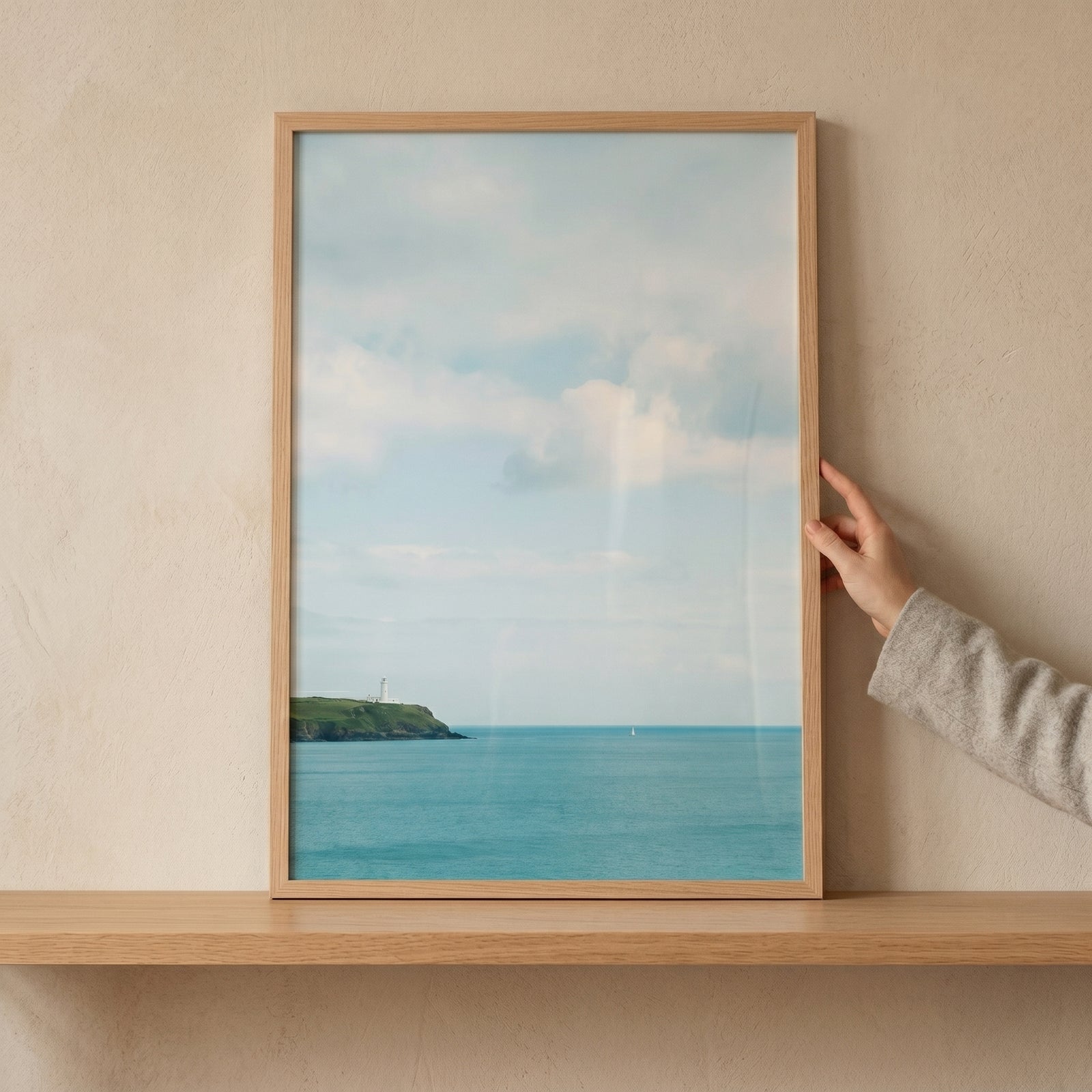 Hand holds wood framed coastal turquoise sea lighthouse wall art on a shelf against a neutral wall.