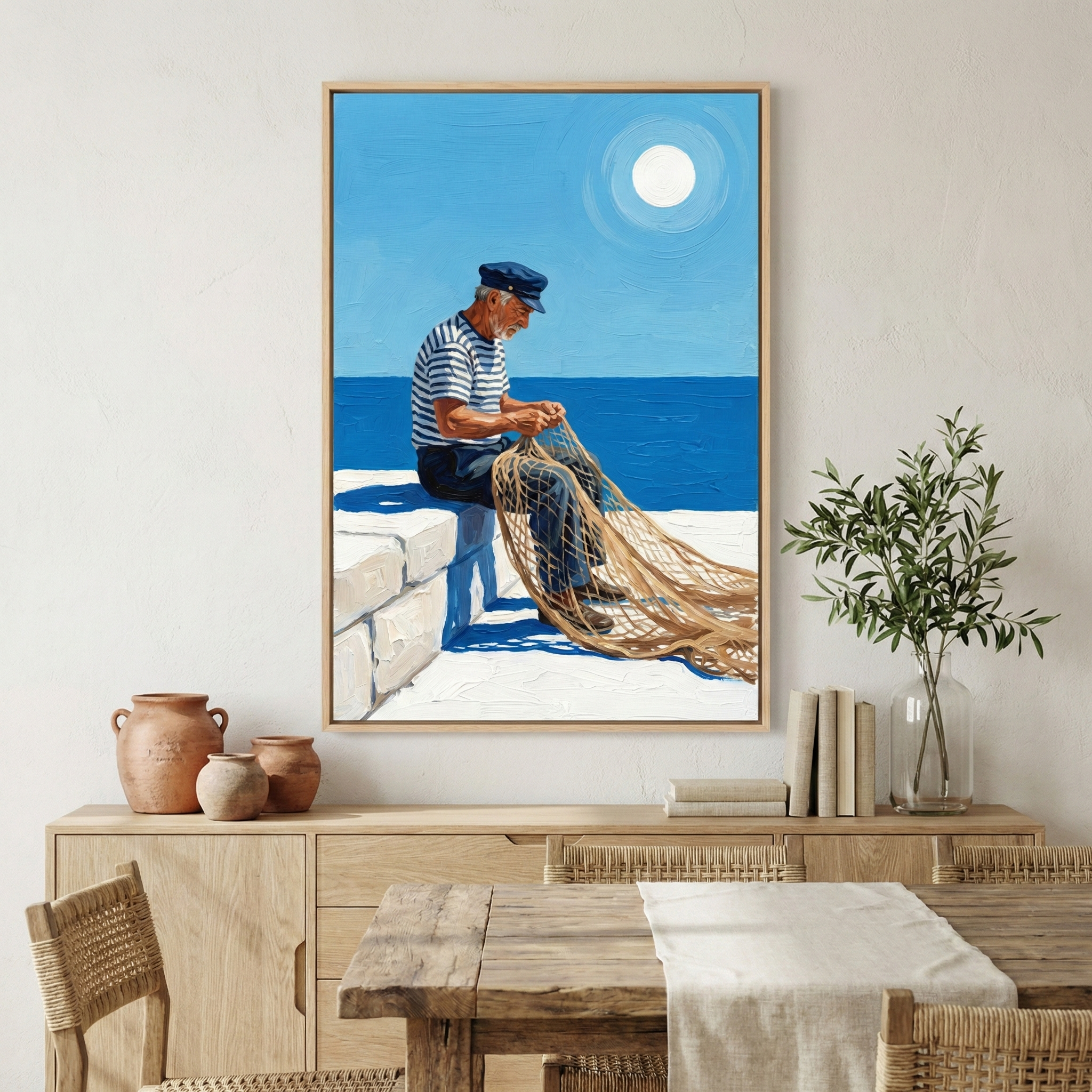 A framed artwork is displayed on a light-colored wall above a wooden sideboard. The central image shows an older man with a white beard, dressed in a blue and white striped shirt and a dark sailor's cap. He is sitting on a bright white stone wall, focused on mending a large, golden-hued fishing net. Behind him is a deep blue sea and a clear, lighter blue sky with a large, brilliant white sun. 
