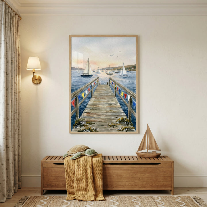 A vertical, light-wood framed artwork is the central focus, hanging on a neutral-colored wall in a bright room. The artwork is a coastal illustration of a long wooden pier adorned with colorful signal flags that extends into a calm harbor filled with white sailboats. 