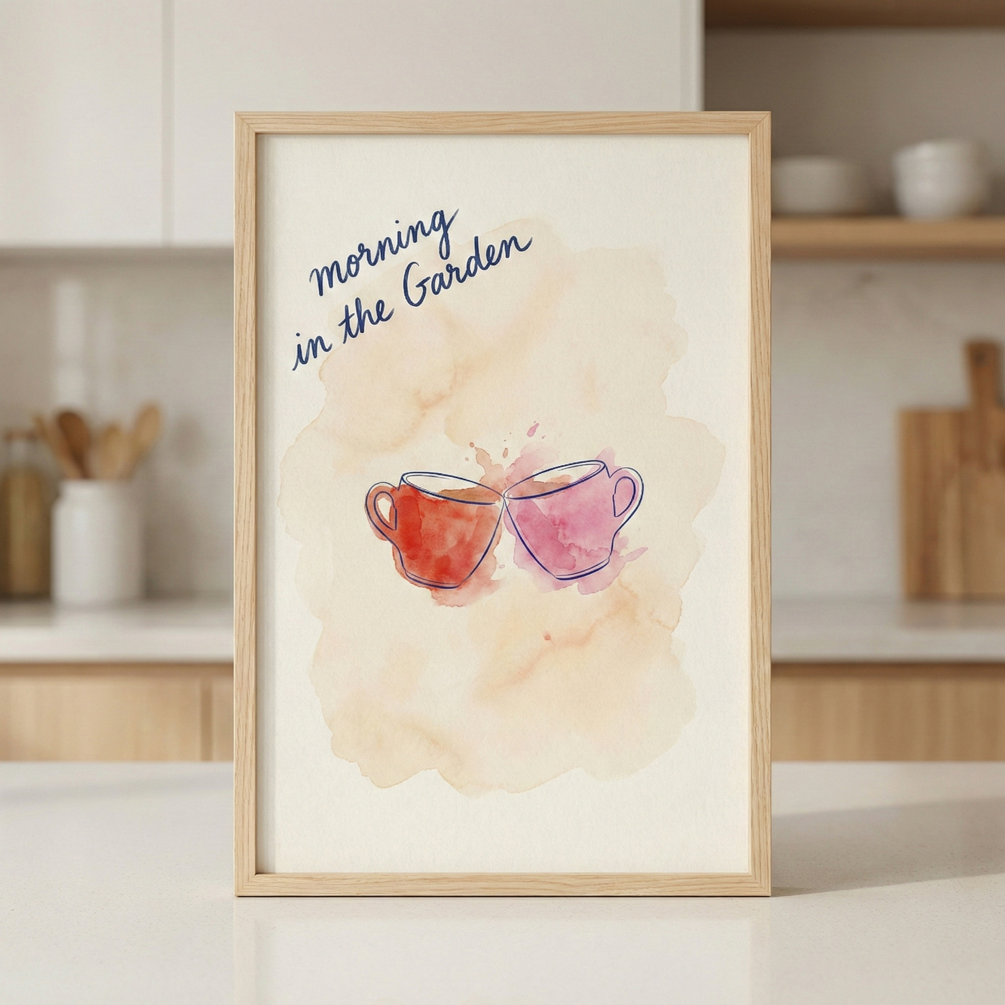 A framed illustration featuring two watercolor-style mugs, one reddish-orange and one pink, clinking together against a soft peach watercolor backdrop. Above the mugs, the text "morning in the Garden" is written in a dark blue script. The piece is housed in a light-colored wood frame and is displayed on a white kitchen countertop with a blurred background of white cabinets and kitchen tools.